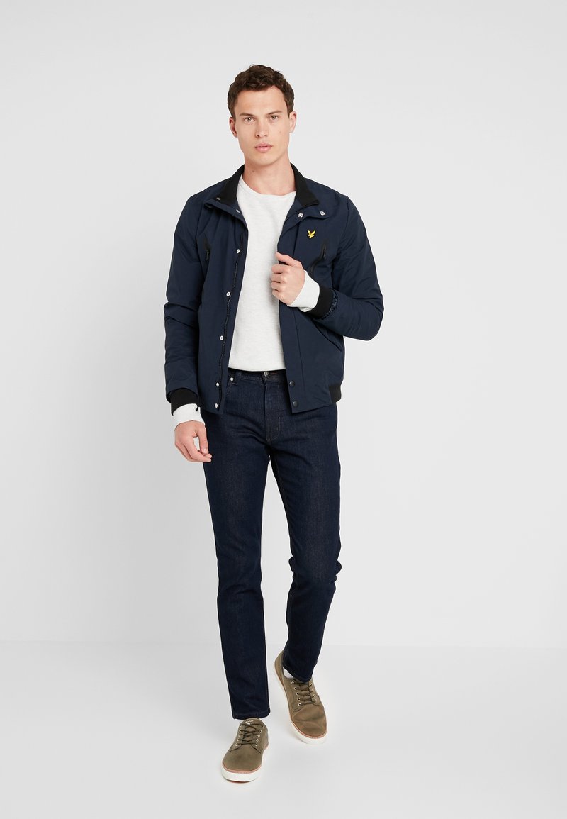 Granatowa lekka kurtka z zamkiem z przodu, dwoma zamkami błyskawicznymi na kieszeniach i małym logo. Noszona na białym swetrze, w połączeniu z ciemnymi dżinsami i oliwkowymi trampkami.