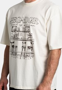 Camiseta de manga corta de color crema con un dibujo en línea negro de una escena de bar, con el texto "PEGADOR" y "Aroma de verano" arriba.