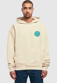 Beige hettegenser laget av mykt stoff, med en frontlomme, snor i hetten og et sirkulært turkist grafikk som sier "DANCE UNDER STARS."