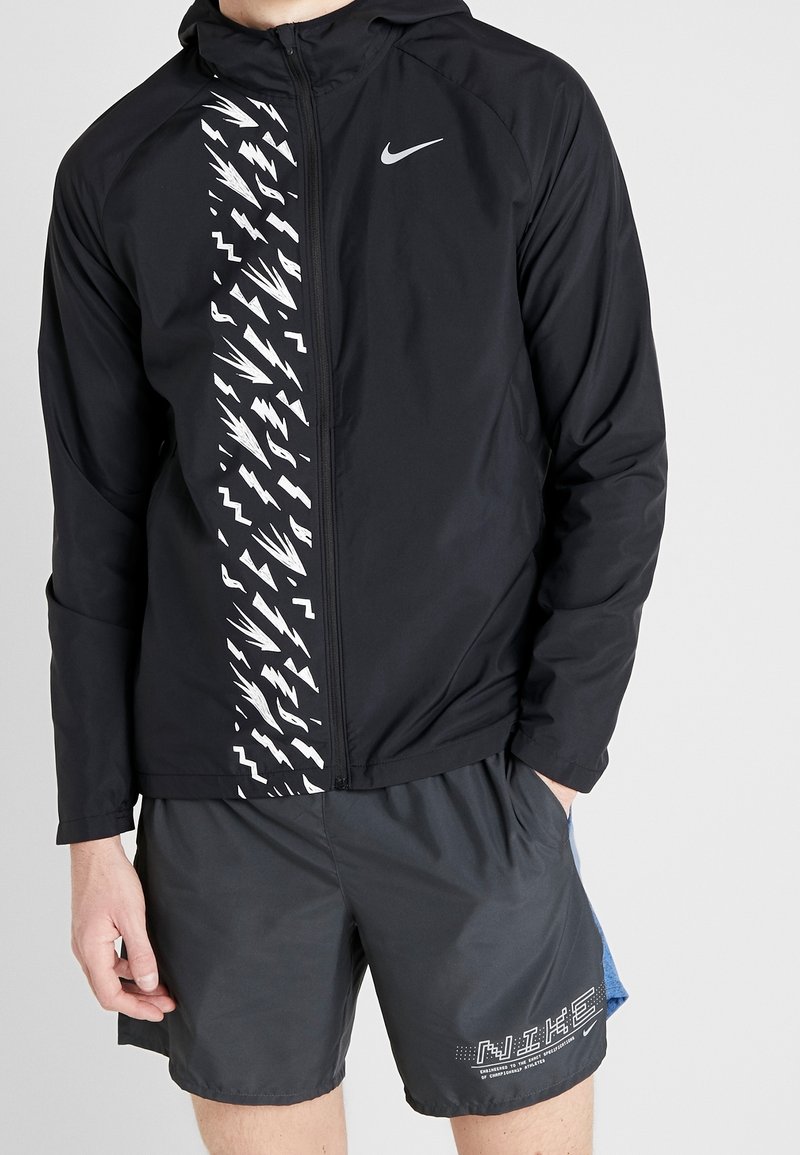 Zwarte lichtgewicht jas met een wit zigzagpatroon aan de voorkant en een Nike-logo. Gekozen in combinatie met grijze shorts met een grafische print.