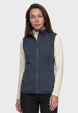 Frau mit langen braunen Haaren, die eine graue Fleece-Weste mit Reißverschluss über einem hellen Langarmshirt und schwarzen Hosen trägt, steht vor einem schlichten Hintergrund.