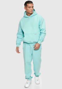 Helles blaues, übergroßes Hoodie mit einer Fronttasche, kombiniert mit passenden Jogginghosen. Beide Teile haben eine glatte Textur und gerippte Bündchen. Weiße Sneakers.