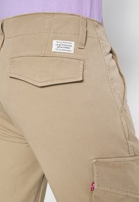 Béžové cargo kalhoty s čtvercovou zadní kapsou, šitím a štítkem s nápisem "Levi Strauss & Co." Červený akcent na švu. Hladká textilní struktura.
