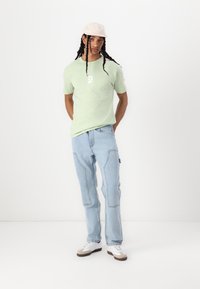 Lichtgroene t-shirt met korte mouwen en een witte graphic, gecombineerd met lichtblauwe jeans met contrasterende naden en een relaxte pasvorm, aangevuld met een roze pet.