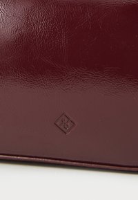 Portefeuille en simili cuir bordeaux avec une finition texturée, présentant un petit logo embossé dans le coin et un design rectangulaire épuré.