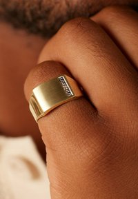 Gouden metalen ring met een geborstelde afwerking en een rechthoekige inset met kleine heldere stenen. Gladde, brede bandontwerp.