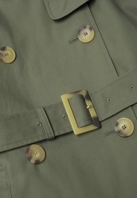 Tissu vert olive avec quatre boutons écaille de tortue marron et boucle de ceinture rectangulaire assortie sur un trench coat.