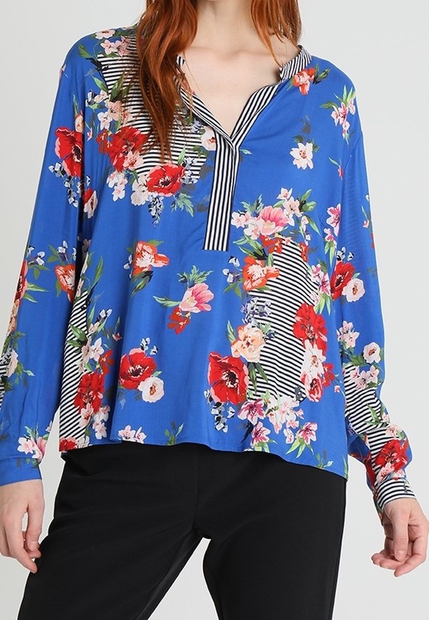 Blusa azzurra a maniche lunghe con stampa floreale rossa e rosa e dettagli a righe in bianco e nero, abbinata a pantaloni neri.