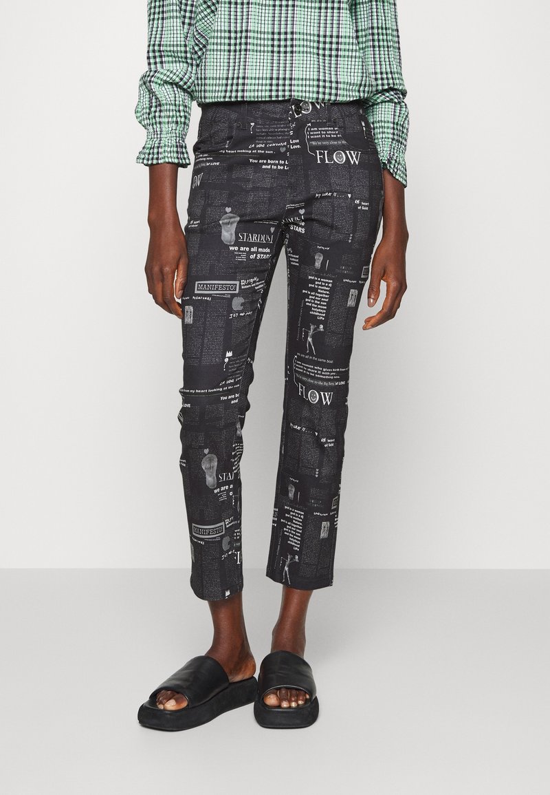 Pantalons noirs avec un design imprimé comportant du texte et des éléments graphiques. Coupe ajustée et longueur cropped, associés à des sandales noires simples.