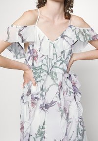 Blommig klänning med volangärmar som lämnar axlarna bar, V-ringning, lagerdesign och genomskinligt tyg med gröna och rosa botaniska mönster.