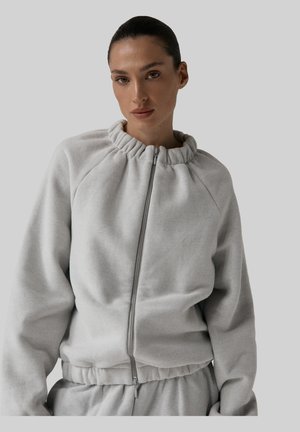 Kobieta z włosami zaczesanymi do tyłu, ubrana w jasnoszary zamek błyskawiczny i dopasowane spodnie, na szarym tle.