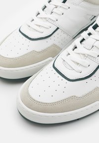 Gros plan de baskets blanches avec orteils perforés, accents en daim beige, bordure foncée et lacets blancs sur fond blanc.