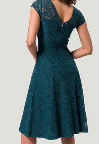 Robe en dentelle teal avec de courtes manches à manches, un corsage ajusté et une jupe évasée. Motif de feuilles notable et détail de fermeture éclair dans le dos.