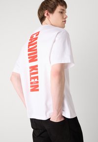 Hvit bomulls T-skjorte med en fet rød "CALVIN KLEIN" logo på ryggen, korte ermer og en avslappet passform. Stylet med svarte bukser.