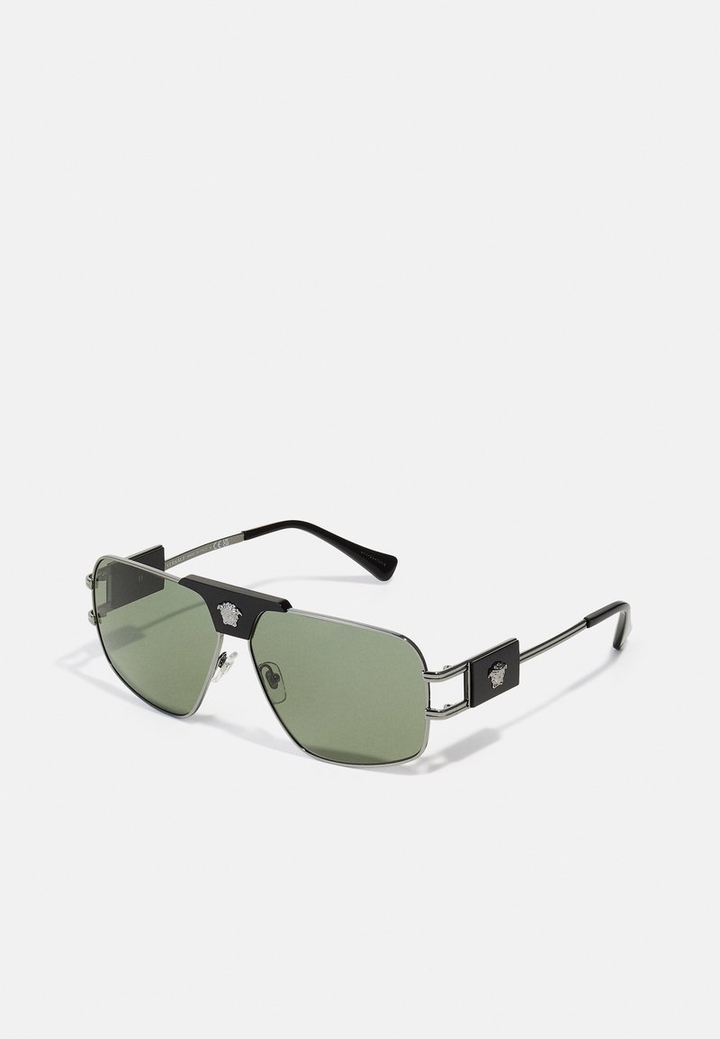 Lunettes de soleil avec des verres teintés en vert, barre supérieure noire, cadre en métal argenté, et accents matériels distinctifs sur les branches.
