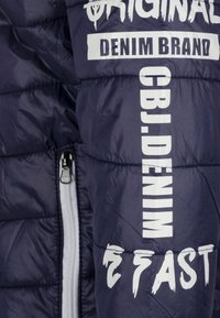 Chaqueta acolchada en azul marino con texto de marca en blanco. Presenta un cierre de cremallera y una textura acolchada. Con detalles de costura visibles.