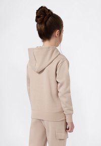 Meisje met bruin haar in een knot, gekleed in een beige hoodie en bijpassende broek met een zijzak, met de rug naar de camera tegen een witte achtergrond.