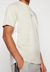 Béžové bavlněné tričko s krátkým rukávem, se vzorovými bílo-černými pruhy na stranách a malým logem na hrudi. Hladká struktura, standardní střih.