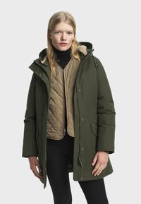 Žena v zelenej zimnej bunde s kapucňou, cez krémovú prešívanú vestu a čierny rolák, postavená proti jednoduchému pozadiu.