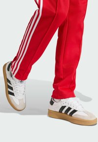 Sapatilhas brancas com detalhes em preto e três riscas, apresentando uma biqueira em camurça e sola de borracha, combinadas com calças vermelhas com riscas brancas.