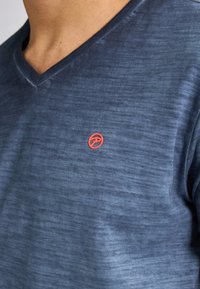 Tričko s výstřihem do V v tmavě modré barvě s texturovaným povrchem. Obsahuje malé červené logo na hrudi. Krátké rukávy, ležérní design.