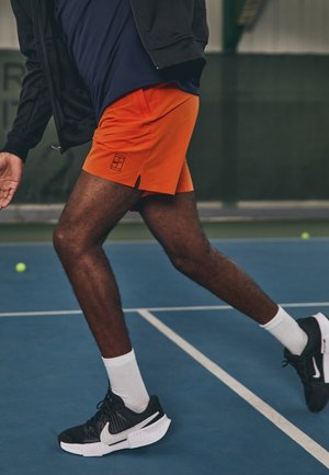 Homme en short orange et chaussures de sport noires sur un court de tennis, se préparant à se déplacer ou à frapper une balle.