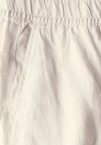 Ljust beige tyg med en mjuk textur, som har ett elastiskt midjeband och veckad design. Sömnadsdetaljer är synliga.