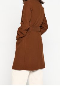 Vrouw draagt een bruine wollen jas met riem en witte broek, gezien van achteren met handen in de jaszakken.