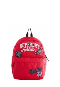 Röd ryggsäck i tyg med rundad form. Har vit broderad text "SUPERDRY Athletics" och olika lappar framtill.
