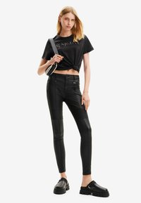 Camiseta negra corta con nudo en el frente y letras metálicas, combinada con leggins de cuero negros que presentan paneles texturizados y cremalleras.