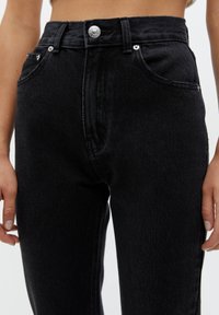 Gros plan sur une personne portant un jean en denim noir taille haute avec des poches avant et une fermeture à bouton métallique.