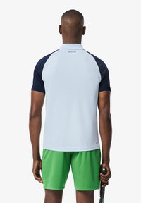 Polo in azzurro chiaro con maniche raglan blu navy, realizzato in tessuto morbido; abbinato a pantaloni corti verdi. Include un logo Lacoste sul retro.