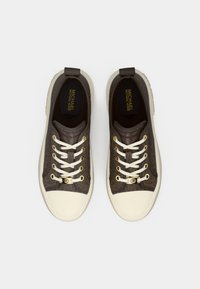 Zapatillas marrones con estampado, punteras y cordones de goma blanca. Presentan detalles dorados y una parte superior de tela marrón con un logotipo.