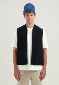 Svart ärmlös väst med texturerad tyg och framdragkedja, kombinerad med en vit t-shirt och beige byxor. Har två framfickor.