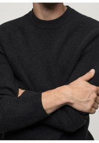 Schwarzer Strickpullover mit rundem Halsausschnitt, strukturiertem Stoff und gerippten Bündchen. Mit verschränkten Armen, die das weiche Material und das figurnahe Design hervorheben.