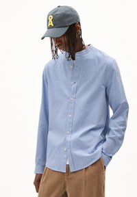 Helles blaues Hemd mit Rundkragen, langen Ärmeln und dezenten vertikalen Streifen, getragen mit einer grauen Kappe mit gelbem Stickereimuster.