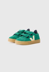 Sapatos verdes em camurça com duas tiras de velcro pretas, logotipo lateral em creme e solas de borracha de goma. Bico arredondado e design baixo.