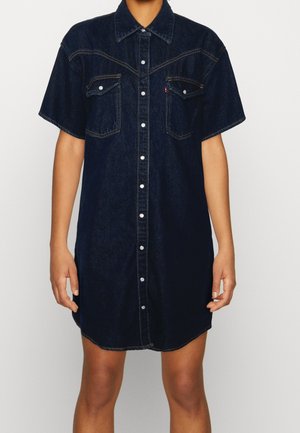 Short-sleeve dark blue denim shirt dress with front snap buttons and two chest pockets, worn by a standing person.