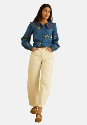 Femme portant un chemisier bleu à broderies florales avec col à volants, pantalon ample beige, ceinture marron et chaussures à brides marron, debout les mains dans les poches.