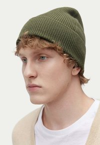 Olivgrön ribbad stickad mössa med lätt slouch, kombinerad med en vit t-shirt och en beige kofta, som har subtil textur och design.