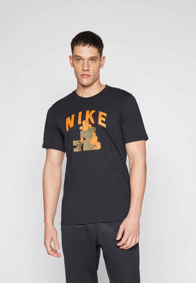 Sort bomulds T-shirt med et orange Nike-logo og illustration. Rund hals, korte ærmer, afslappet pasform. Båret med grå shorts.