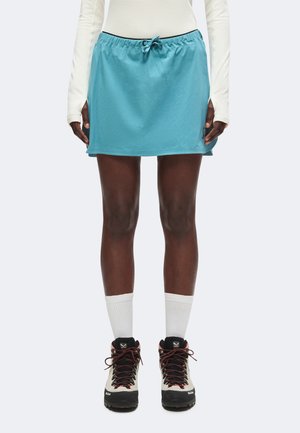 Person trägt einen hellblauen Rock mit Kordelzug, ein weißes Langarmshirt, weiße Socken und schwarze Wanderschuhe und steht aufrecht.