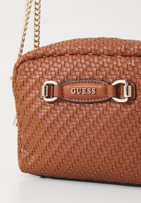 Tkanasta smeđa torba napravljena od umjetne kože, s zlatnom lancem i označenom kožnom oznakom s uklesanim natpisom "GUESS".