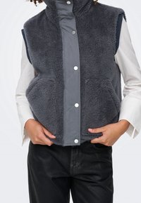 Personne portant un gilet en polaire gris foncé avec des boutons-pression sur une chemise blanche à manches longues et un pantalon noir, les mains reposant près des poches du gilet.