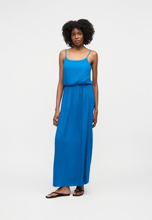 Nainen seisoo sininen hihaton, ohuilla olkaimilla varustettu maksimekko päällään ja mustat varvassandaalit jalassa yksinkertaisella vaalealla taustalla.