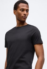 Tânăr privind lateral, purtând un tricou simplu negru cu mânecă scurtă, pe un fundal deschis simplu.