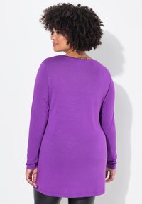Lila Langarm-Tunika mit Rundhalsausschnitt, weicher Textur und lässiger Passform, die bis zur Mitte des Oberschenkels reicht, getragen über schwarzen Leggings.