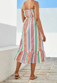 Robe maxi sans bretelles avec des rayures multicolores, présentant des motifs floraux et géométriques, un ourlet évasé et une taille élastique. Sandales marron visibles.