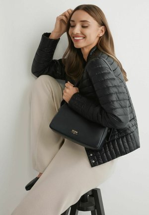 Uśmiechnięta kobieta siedząca na stołku, ubrana w czarną pikowaną kurtkę i kremowe spodnie, trzymająca czarną torebkę Céline na białym tle.