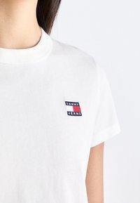 Vit bomullströja med rund halsringning. Har en liten, flerfärgad "TOMMY JEANS" logotyp på vänster br öst. Korta ärmar.